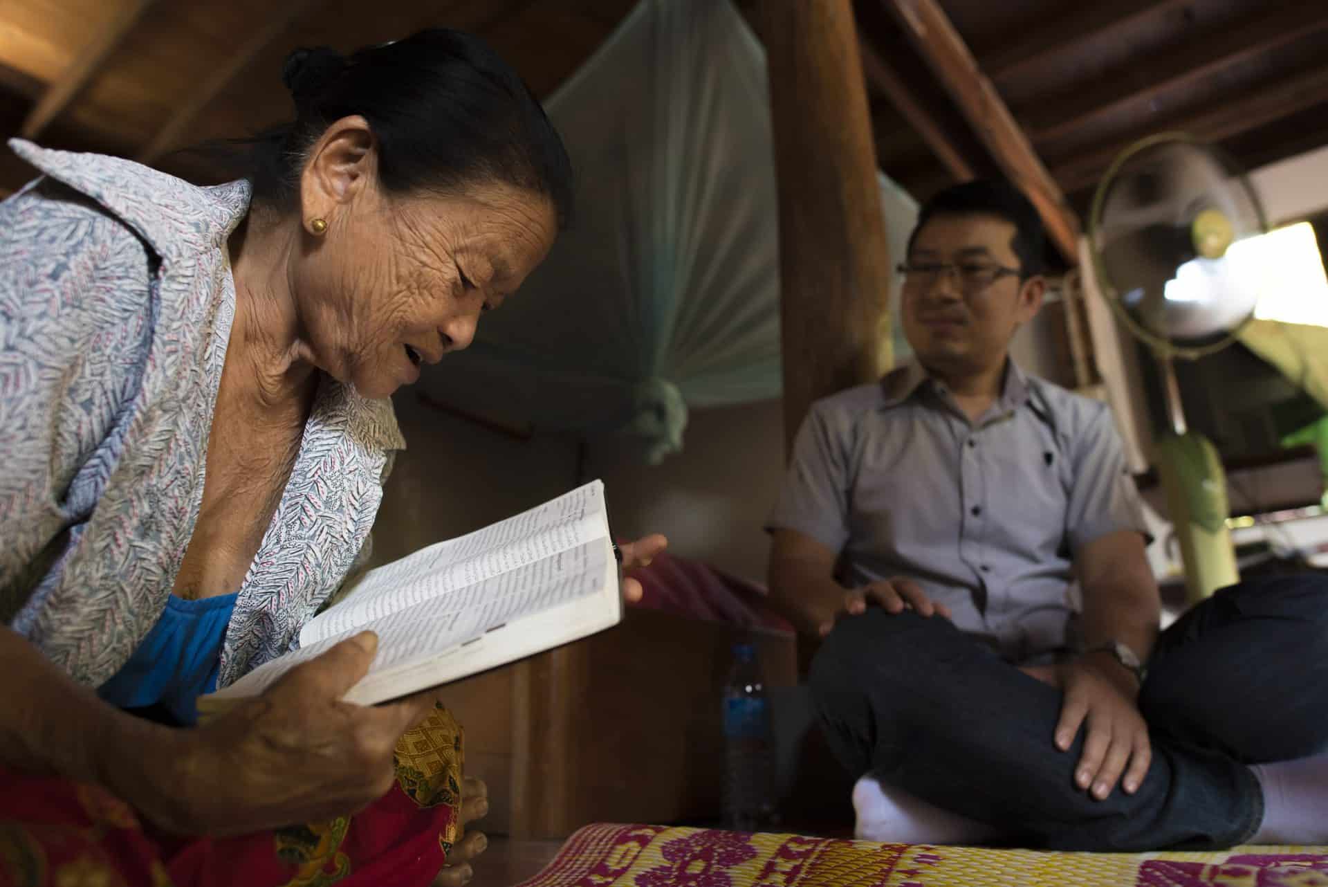 A woman reads the bible while a younger man looks on.